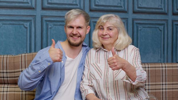 Joyful bonding loving generations family of senior mother and handsome adult son or grandson showing thumbs up gesture. Cheerful excited young man with gray-haired old mature mom or grandmother