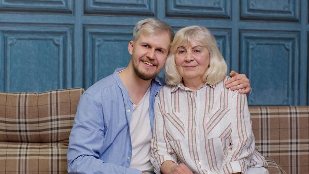 Cheerful young man with gray-haired old mature mom or grandmother. Domestic weekends. Bonding loving generations family of senior elderly mother and handsome adult son or grandson looking at camera.