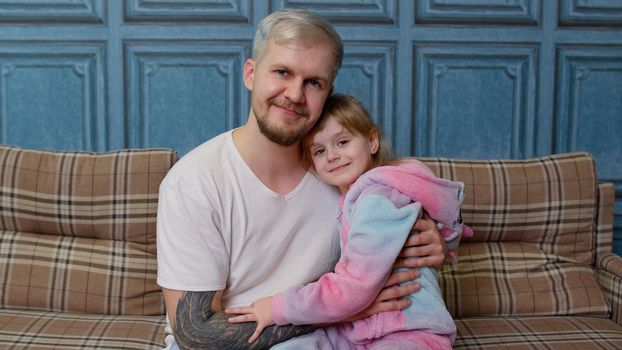 Father and little child daughter kid in pajamas sit on couch in living room smiling, looking at camera, having fun, embracing together indoors. Girl hugging young adult man. Bonding family generation