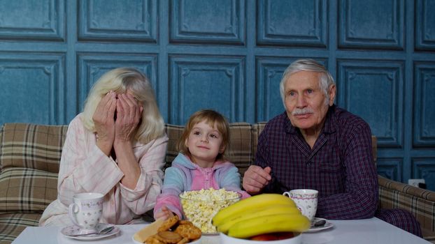 Grandparents watch TV with granddaughter at home. Senior grandfather and grandmother, using family time relax with young girl kid sitting on sofa in living room getting scared watching horror movie