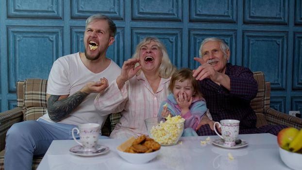 Multigenerational happy family laughing, watching cartoons television movies eating popcorn at home. Senior couple grandfather and grandmother relaxing with adult son and little girl kid granddaughter