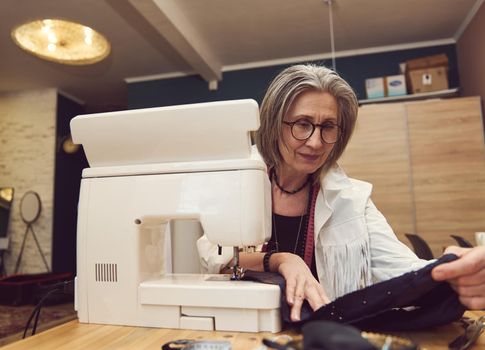 Mature Caucasian woman seamstress works, sews clothes on a sewing machine. The process of sewing in an atelier or workshop. Tailoring and repair of clothing. Self-employed employee.