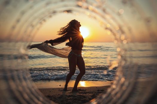 A beautiful creative composition of a sea landscape shot through a circle focus showing a young woman who is dancing on the beach in sunset.