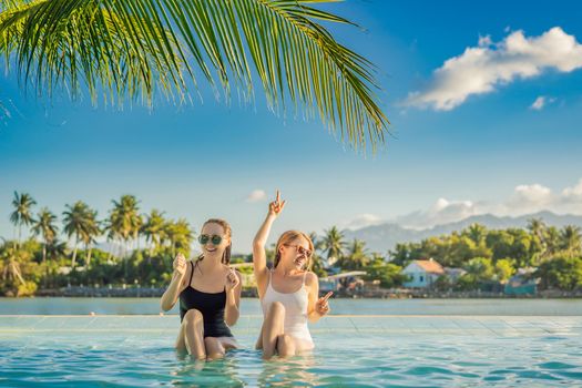 Two beautiful young woman sitting by the poolside of a resort swimming pool during summer holiday. Party, tourism in tropical countries.