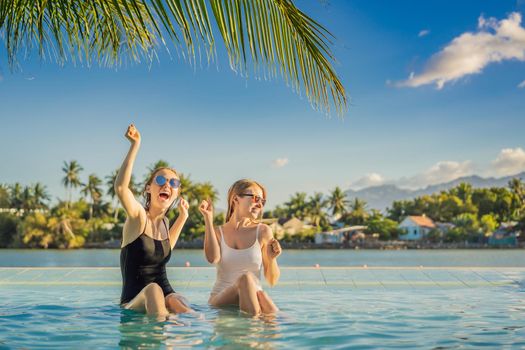 Two beautiful young woman sitting by the poolside of a resort swimming pool during summer holiday. Party, tourism in tropical countries.