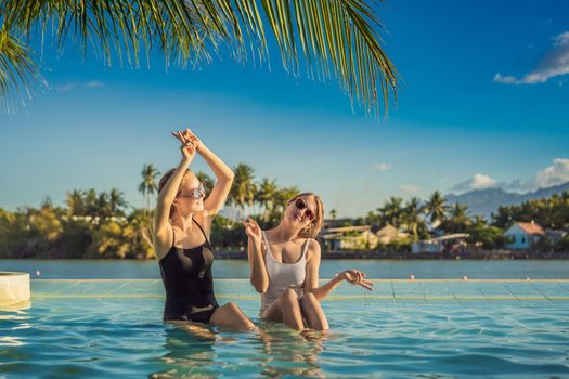 Two beautiful young woman sitting by the poolside of a resort swimming pool during summer holiday. Party, tourism in tropical countries.