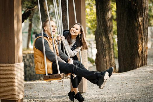 Side view of young loving couple sitting on swing in park outdoors. High quality photo