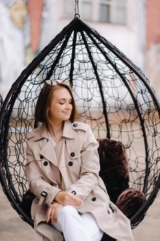A happy stylish girl In a gray coat is sitting outside in an armchair.
