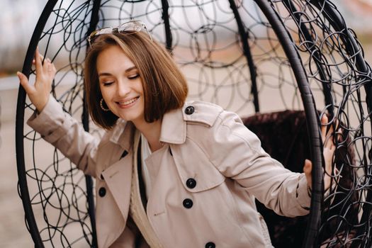 A happy stylish girl In a gray coat is sitting outside in an armchair.