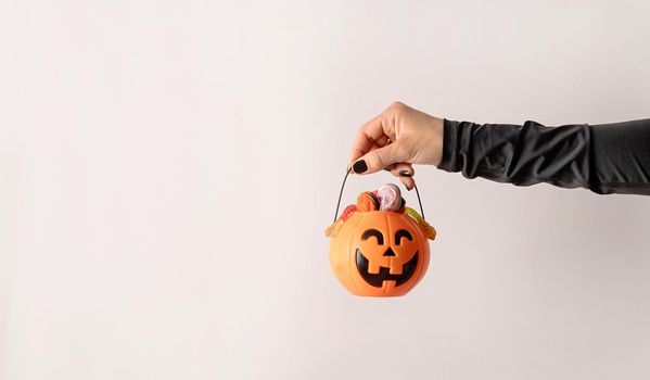 Happy Halloween concept. Scary woman Halloween hand with black nails holding pumpkin full of sweets on white background with copy space