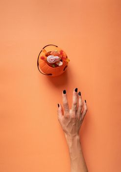 Happy Halloween concept. Scary woman hand with black nails trying to get pumpkin with sweets, flat lay on orange background