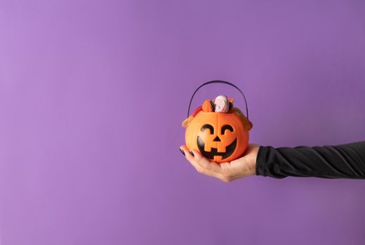 Happy Halloween concept. Scary woman Halloween hand with black nails holding pumpkin full of sweets on purple background with copy space