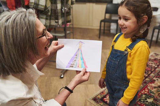 Pleasant elderly Caucasian woman tailor, caring and loving grandmother smiles admiring the clothes sketch of her adorable granddaughter while working in a fashion design studio and tailoring atelier