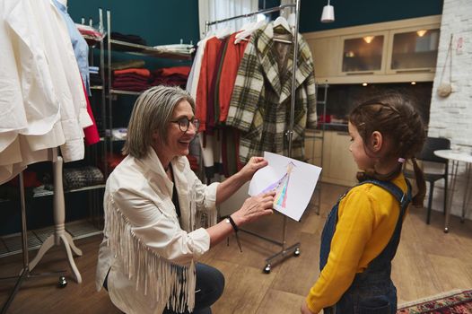 Pleasant mature European woman, cheerful caring and loving grandma admiring the drawing - clothes sketch- of her granddaughter adorable baby girl while working in a fashion design workshop