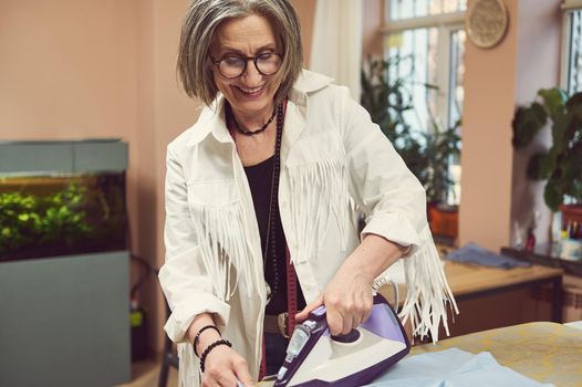 Pleasant charming mature Caucasian woman seamstress tailor strokes the blue shirt from new collection in a sewing workshop. Sewing process, fashion design, creation of new garment in tailoring atelier