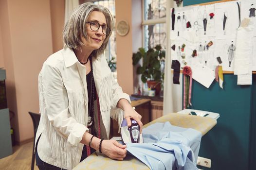 Pleasant charming mature European woman seamstress tailor strokes the blue shirt from new collection in a sewing workshop. Sewing process, fashion design, creation of new garment in tailoring atelier