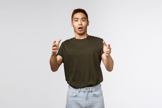 Portrait of shocked young male, taiwanese guy catching product that person throwing at him, raising hands up and look intense and stressed, gasping startled, standing grey background.