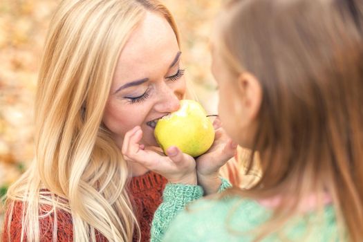 Portrait of little girl with young mother eating yellow apple in autumn park