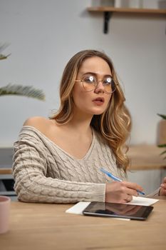 Blonde female blogger in glasses, beige knitted sweater. Sitting in kitchen at wooden table with pink cup, palm tree and tablet on it. Going to write something in notebook. Work or education. Close-up