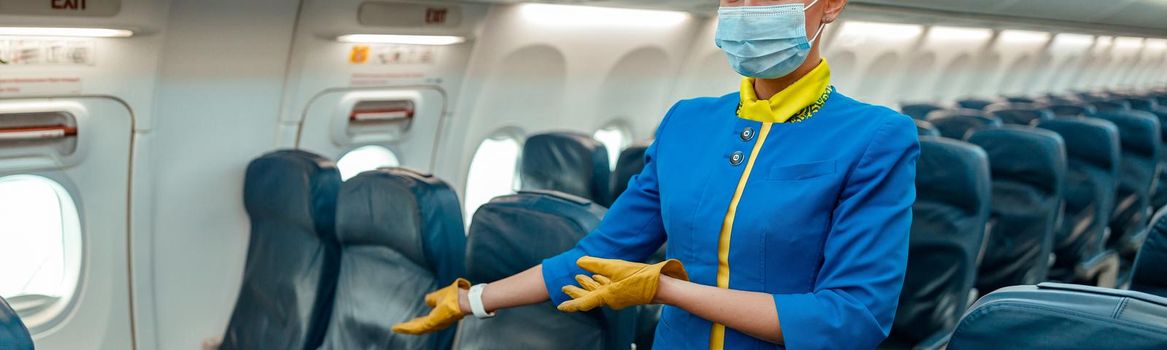 Woman flight attendant wearing protective medical mask and air hostess uniform while gesturing towards passenger chair in airplane