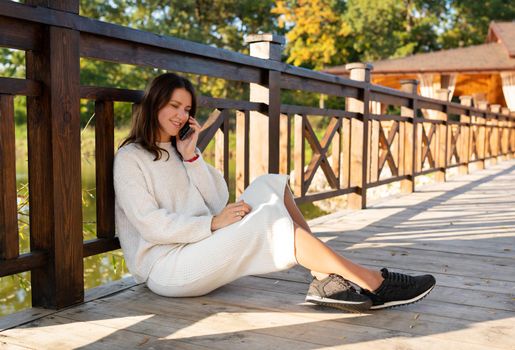 The girl is talking on the phone while sitting on the street on a wooden bridge. A beautiful sunny fall day