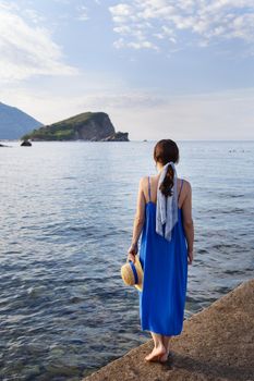 Montenegro, Budva. Girl in a blue dress holding a straw hat, she stands on a wooden pier by the sea, beautiful sky with clouds. Vacation concept, travel to Europe