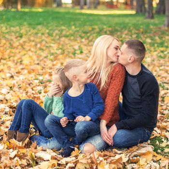 Portrait of young family sitting in autumn leaves. Parents kissing and sitting with children in the autumn park