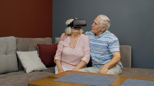 Senior wife playing game in VR headset glasses, husband laughing with her action, elderly couple lifestyle concept. Family of senior man and woman at home. Future technology. Virtual reality goggles