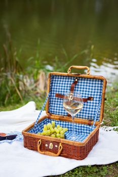 Romantic picnic in the park on the grass against the backdrop of a beautiful lake, delicious food: picnic basket, wine, grapes, figs, cheese, blue checkered tablecloth, two glasses of wine.Outdoor recreation concept
