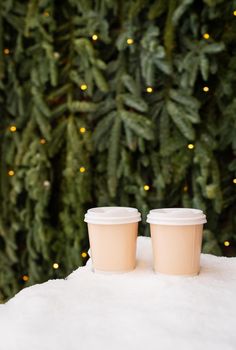 Two paper coffee cups outdoors in the snow against the backdrop of a beautiful green nobilis wall, coffee to go concept. Walks in the open air