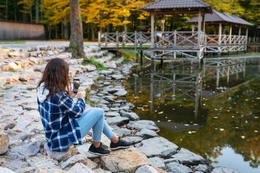 Woman traveler in a plaid shirt makes a photo of the lake with a mobile phone. Beautiful autumn forest