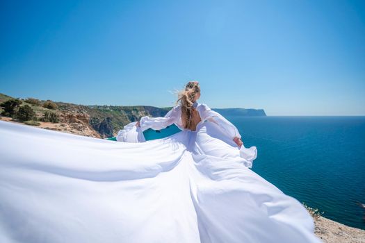 Blonde with long hair on a sunny seashore in a white flowing dress, rear view, silk fabric waving in the wind. Against the backdrop of the blue sky and mountains on the seashore