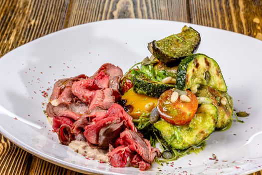 Sliced rare roast sirloin of beef with roasted vegetables on rustic wooden background.