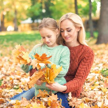 Happy young caucasian woman and little girl holding autumn yellow leaves sitting at the park