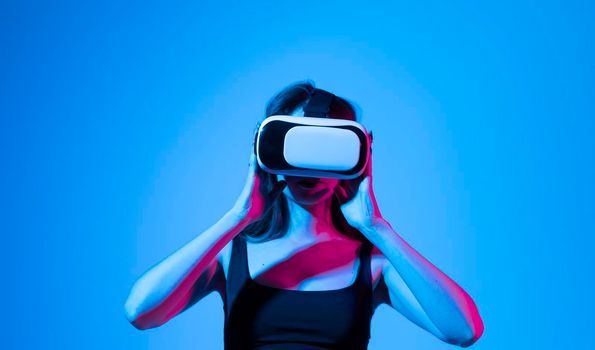 Concept of modern gadgets and technologies. Future technology concept. Portrait of young woman in VR-glasses in neon light on blue background. Virtual reality gaming