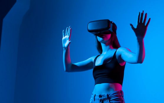 Cheerful girl with hands up wearing the virtual reality goggles and playing a games in neon light. Future technology concept. Metaverse