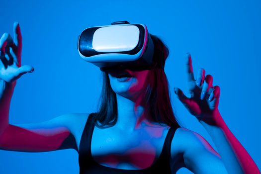 Amazed young woman touching the air during the VR experience. Girl uses a virtual reality headset in neon light. The woman with glasses of virtual reality