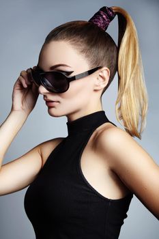 Beautiful young woman with black fashion sunglasses and glamour ponytail hairstyle on blue background. Trendy look for lady. Eye wear style.