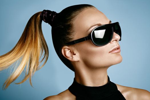 Beautiful young woman with black fashion sunglasses and glamour ponytail hairstyle on blue background. Trendy look for lady. Eye wear style.