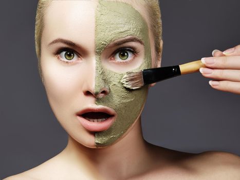 Beautiful Woman Applying Green Facial Mask. Beauty Treatments. Close-up Portrait of Spa Girl Apply Clay Facial mask with brush on grey background