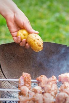 Hands of man prepares barbecue meat with potatoes on skewer by grill on fire outdoors. Concept of lifestyle rustic food preparation