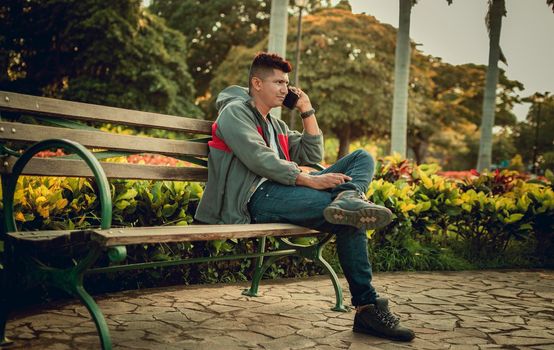 Man sitting on a bench calling on the phone, young man sitting on a bench with cell phone, close up of a young man calling on the phone sitting on a bench