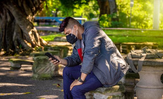 Man with a mask sitting on a bench with his phone, young man with a mask sitting with a cell phone, near a young man with a mask texting