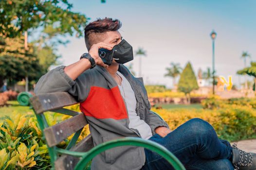 Man sitting on a bench calling on the phone, young man with a mask calling on the cell phone, man with a mask calling on the phone in a park