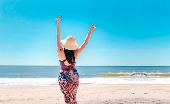 woman with hat from back on the beach, young woman raising her hands on the beach