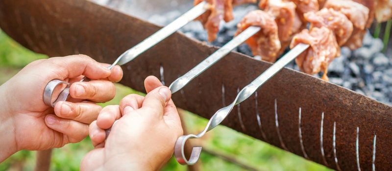 Hands of man prepares barbecue meat on skewer by grill on fire outdoors