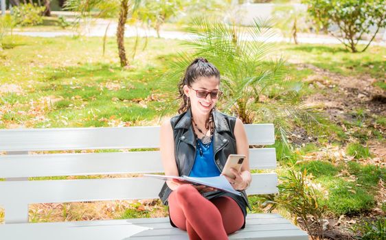 A girl sitting taking notes with a notebook and cell phone, Woman sitting taking notes with her cell phone, Latin girl using her cell phone in a park