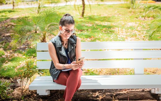 Girl sitting on a bench checking her cell phone, Happy woman sitting in a park texting on her cell phone, Woman sitting on a bench sending a text message