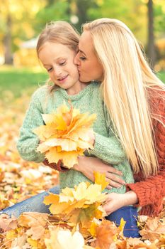 Happy young caucasian mother and little daughter holding autumn yellow leaves sitting and kissing at the park