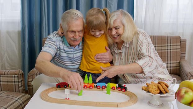 Senior couple grandparents with child kid granddaughter spending time home together, sitting, playing game, riding toy train on wooden railway on table. Old grandmother grandfather with girl grandkid